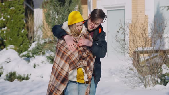 Romantic Loving Young Man Wrapping Smiling Woman with Blanket Standing Outdoors on Sunny Winter Day