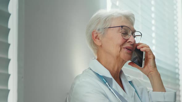 Positive grey haired mature lady doctor with glasses talks on mobile phone