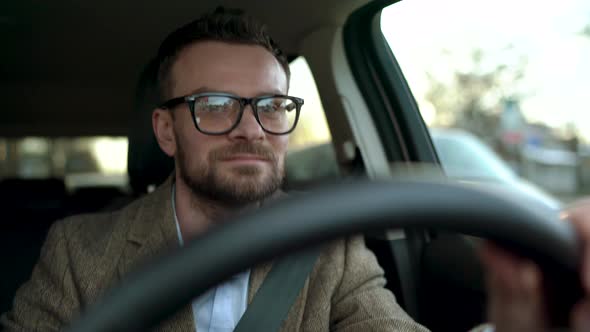 Satisfied Bearded Man in Glasses Driving a Car Down the Street in Sunny Weather