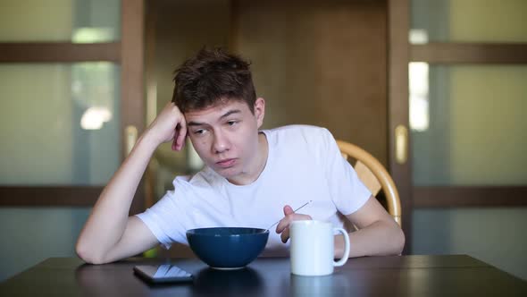 A sleepy teenager lazily eats porridge early in the morning