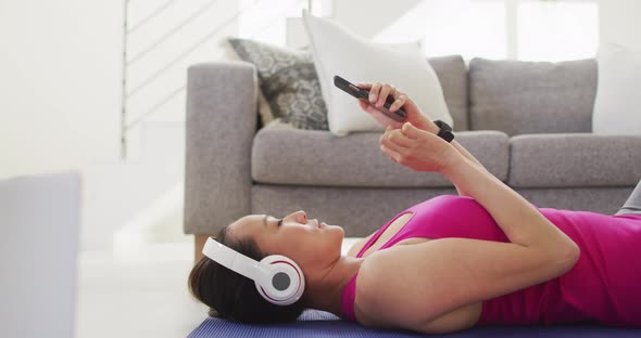 Asian woman in fitness clothes lying on mat wearing headphones, using smartphone at home
