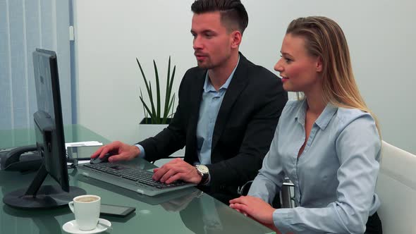 A Man and a Woman Sit at a Desk in Office, He Works on a Computer, She Talks, Then Smiles To Camera