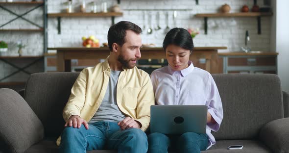 Happy Multiracial Young Couple Talking and Using Laptop Together Watching Video Sitting on Sofa in
