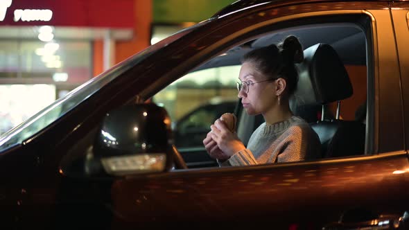 A young woman sits in the car in the evening and eats a burger. Snack on the go