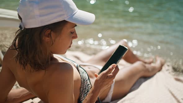 Rear View Relaxed Woman Tourist in Bikini Sunbathes on Sea Beach Close to Water Unlocks Her Mobile