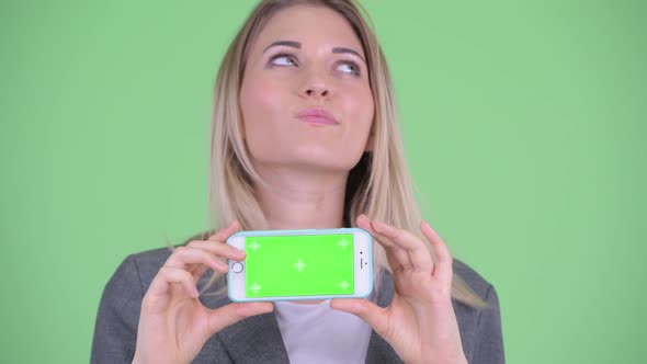 Face of Happy Young Blonde Businesswoman Thinking While Showing Phone