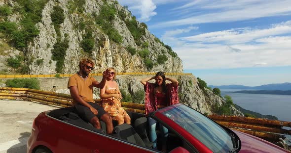 Multi ethnic friends partying in red convertible parked on side of coastal road