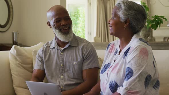 African american senior couple looking at each other and smiling while using laptop together at home