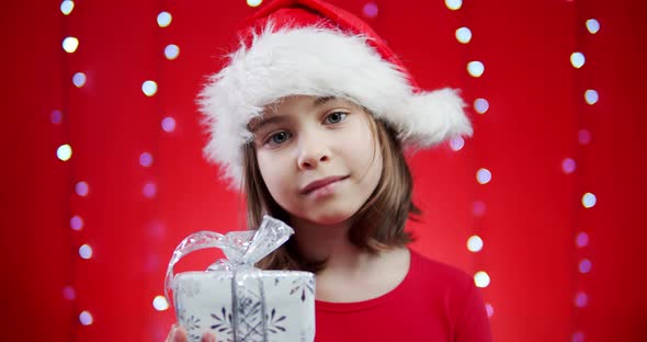 Cute Girl in a Santa Hat Holds a Gift on a Red Christmas Background