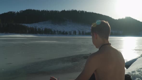 Young man with no shirt sitting beside frozen mountain lake in winter with snow on landscape and bri