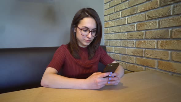 A Young Woman with a Phone Sits at a Table, A Woman Sits at Home at a Table with a Phone