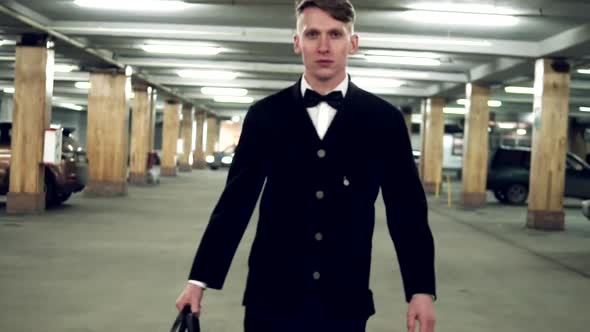 Young Blusinessman in a Black Suit with Bowtie Walking on the Parking with Big Leather Bag