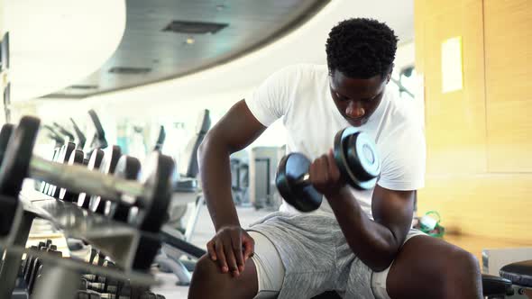 Young African American Man Sitting and Lifting a Dumbbell Close to the Rack at Gym