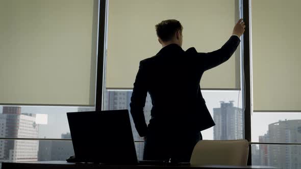 Businessman In Office Standing At Window And Opens The Blinds To Make Brighter