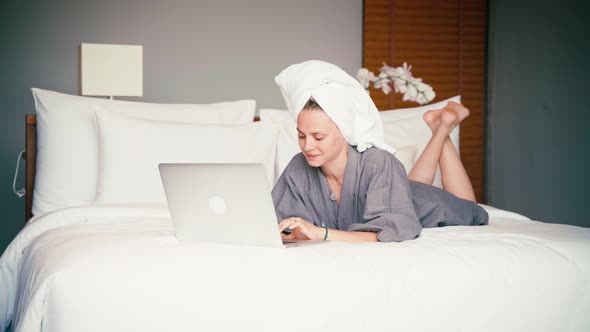 A Woman in a Dressing Gown and a Towel on Her Head Looks at the Laptop Screen