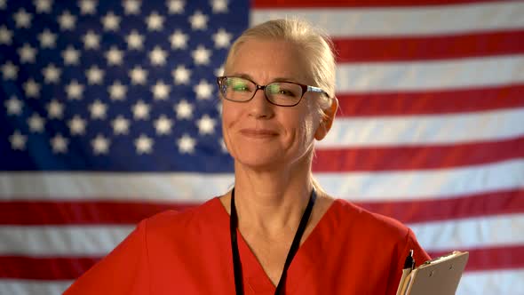 Medium tight portrait of a smiling healthcare nurse with clipboard with out of focus American flag.