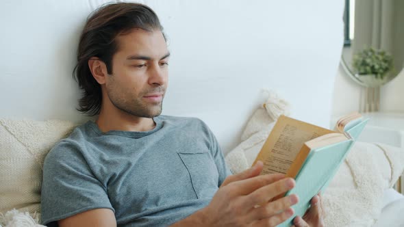 Intelligent Guy in Pajamas Reading Book Smiling Turning Pages Relaxing in Bed at Home