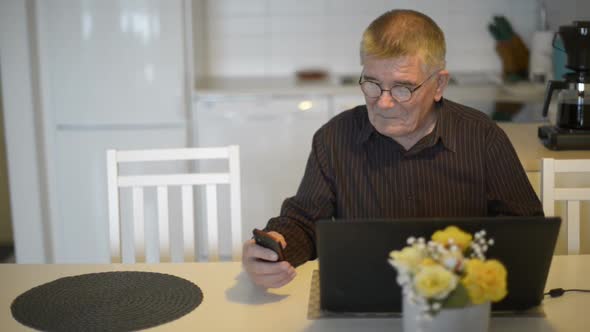 Senior Man with Eyeglasses Using Phone and Laptop in the Dining Room