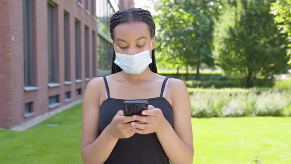 A Young Black Woman in a Face Mask Works on a Smartphone - an Office Building