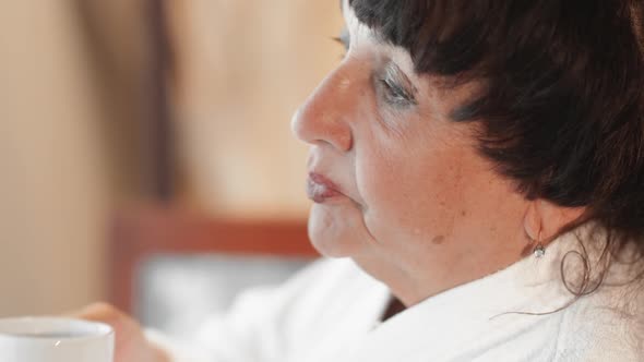 Caucasian Senior Woman Drinking Coffee in the Morning Near a Large Panoramic Window in a White Terry