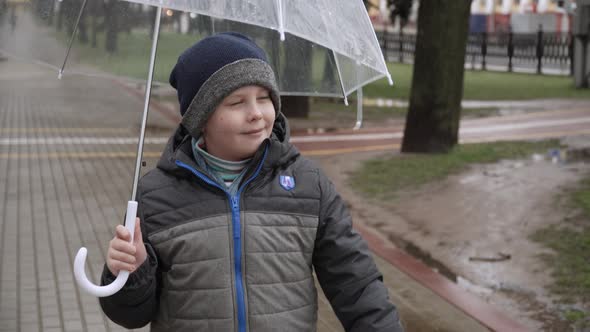 Boy in Hat and Jacket with Transparent Umbrella Walks and Smiles in City Park