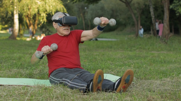 Senior Old Grandfather Man in VR Headset Helmet Making Fitness Exercises with Dumbbells in Park