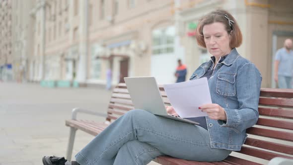 Old Woman Reading Documents and Working on Laptop While Sitting on Bench Outdoor