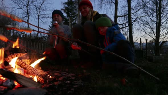 Two boys and their mother sitting around a bonfire with stick bread