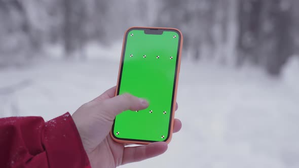 The Smartphone in a Man's Hand Snowy Forest in the Background Fingers Move on the Green Screen of