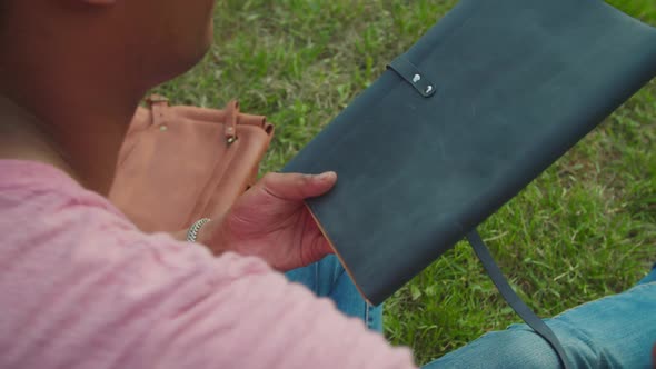 Young Man Opening Leather Sketchbook Sitting on Campus Lawn Outdoors