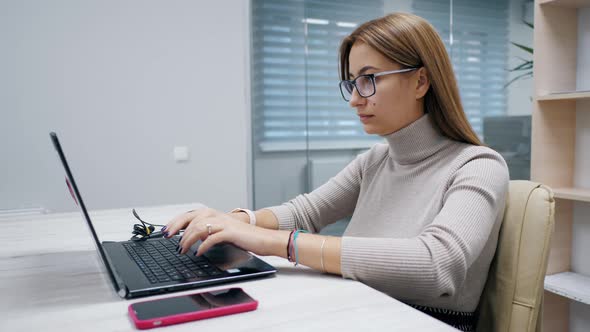 Cute Woman Working at a Laptop with Glasses Typing Text in the Office Employee at Work on the