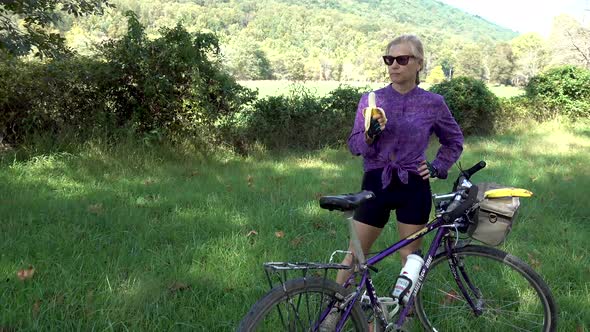 Pretty, blonde, mature woman with sunglasses and gloves next to her bicycle eating a banana on the s