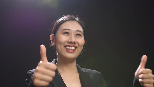 Smiling Asian Speaker Woman In Business Suit Showing Thumbs Up Gesture In The Black Studio