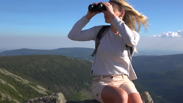 A Young Beautiful Woman Sits on a Rock on a Hilltop and Looks Around Through Binoculars