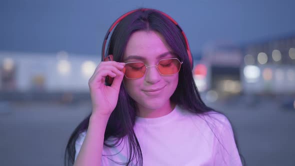 Portrait of Beautiful Young Woman in Sunglasses and Headphones Looking at Camera and Smiling. Close