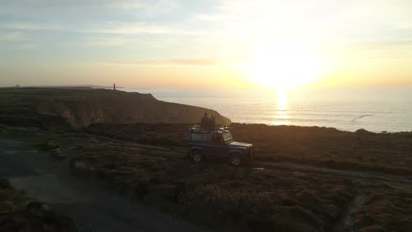 Cinematic aerial of cute couple four by four vehicle along ocean cliffs at beautiful sunset