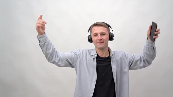 Young Man on a White Background in Headphones and a Smartphone Listens to Music and Dances