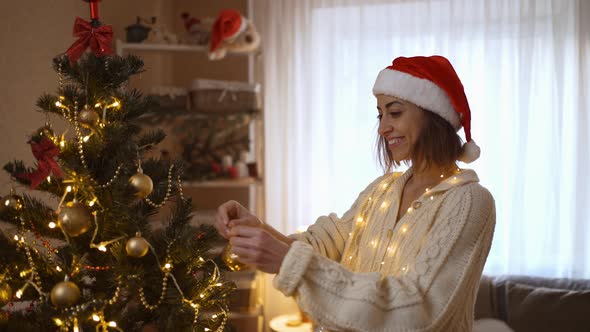 Slow Motion Portrait p of Happy Woman in Santa Hat Hangs Up Golden Ball on Branch of Christmas Tree