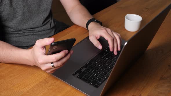 Young hipster with smartwatch typing on laptop and scrolling on smartphone.