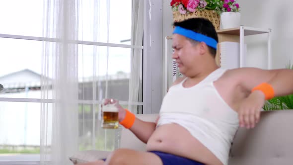 Funny fat Asian guy in sportswear exercise weight training by dumbbell and enjoy drinking beer