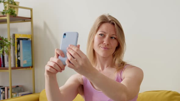 Young Cheerful Blonde Woman in Pink Top Sits on Couch at Home and Takes Selfie on Her Blue Phone