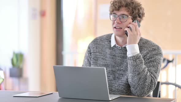 Young Man with Laptop Talking on Smartphone at Work