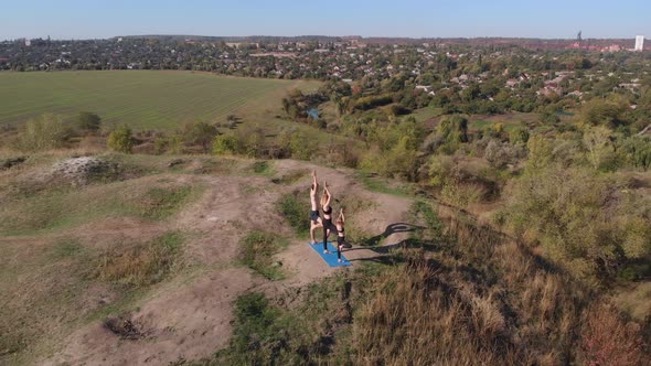Orbiting Aerial Shot of Family of Three Mother Father and Daughter Do Yoga Exercises on Top of Hill