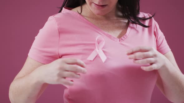 Mid section of a woman touching the pink ribbon on her chest against pink background