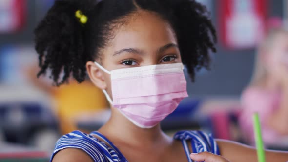 Portrait of african american schoolgirl wearing face mask, sitting in classroom looking at camera