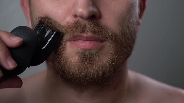 A man shaves his beard and mustache with an electric razor at home