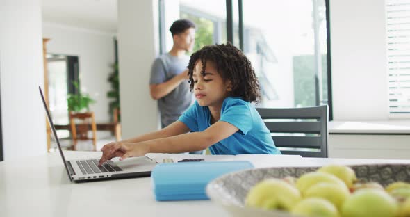 Happy biracial boy sitting at table in kitchen using laptop and doing homework