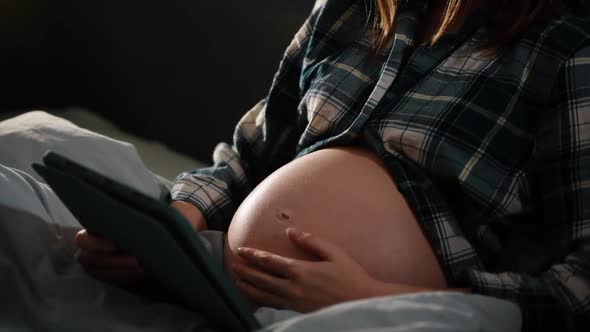 Pregnant with tablet computer in bedroom showing and touching belly.