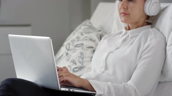 Mid-Aged Woman in Wireless Headphones Using Laptop on Sofa