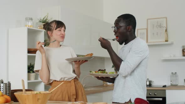 Multiethnic Couple Eating Salad and Chatting in Kitchen